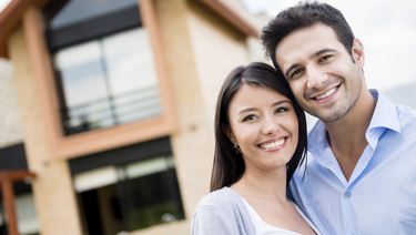 Smiling couple with arms around one another in front of house.