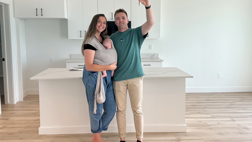 A young family with a baby stands in the kitchen of their new home, holding up their house key