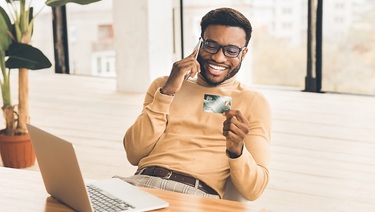 A diverse man talks on his cell phone while holding up a business credit card