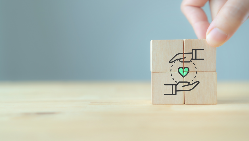 A hand assembling wooden blocks with an illustration of hands surrounding a heart, symbolizing care, support, and compassion