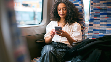 A woman sitting on a train, holding and using a smartphone. The background shows a blurred view of the train window and urban scenery.