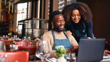 A smiling couple working together in their restaurant, with the man wearing an apron and using a laptop, while the woman leans over to collaborate, surrounded by a warm, inviting atmosphere	