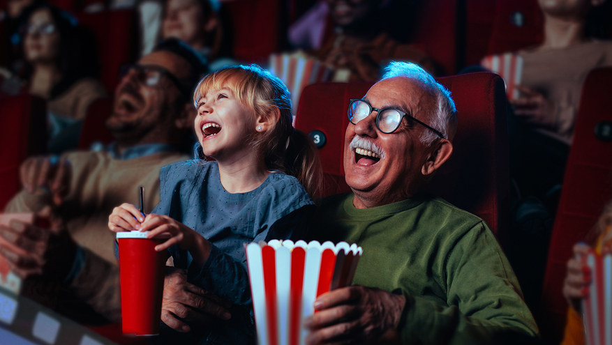 An elderly man with glasses laughs with his young grandchild in a movie theater, both holding popcorn as they enjoy the film