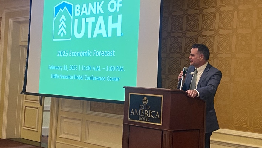 Branden Hansen, president of Bank of Utah, stands behind a wooden podium at the Little America Hotel, speaking into a microphone at the bank’s 2025 Economic Forecast event. 