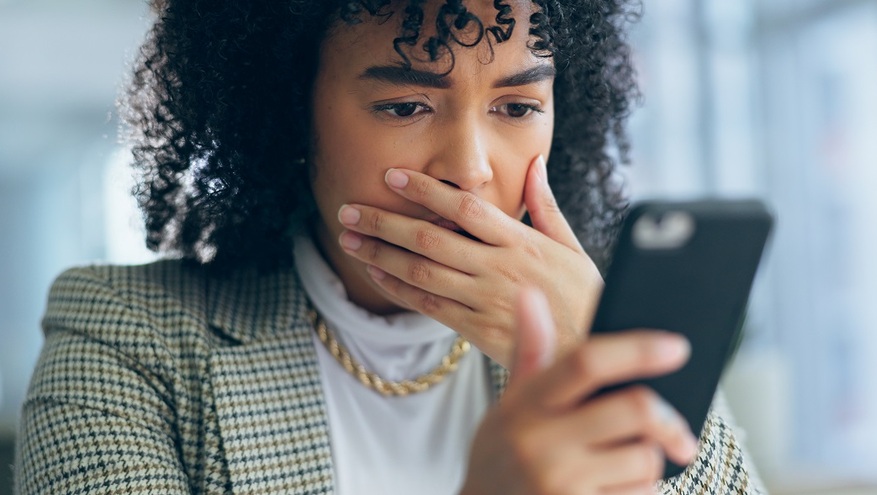 Concerned woman in a checkered blazer looks at her phone, covering her mouth in shock