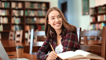 Smiling college student studying in a library with laptop, books and coffee — Bank of Utah, in partnership with College Ave, offers student loans