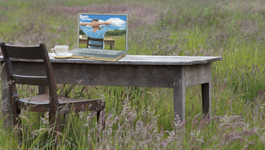 Old barn wood desk with chair in the middle of tall grass field with laptop on the desk. Laptop pictures a man sitting at the same desk in the same field relaxing.
