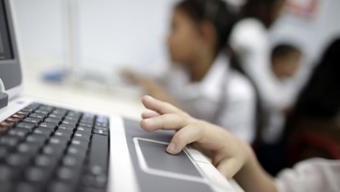 Close up of child's hand on laptop trackpad with other children on computer in blurred background.