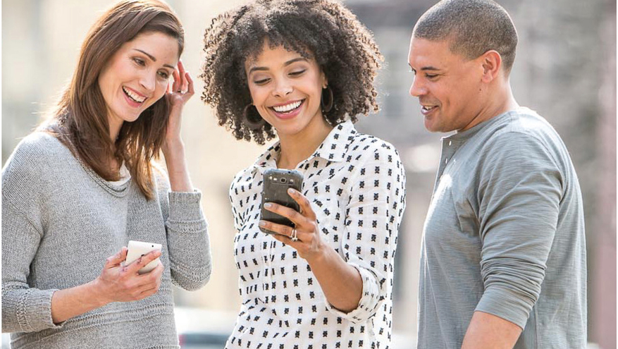 Three young adults smiling and looking at smartphone being help by woman standing in the middle.