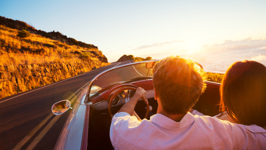 Photo of couple in classic sportscar convertible driving on a scenic, winding road at sunset.