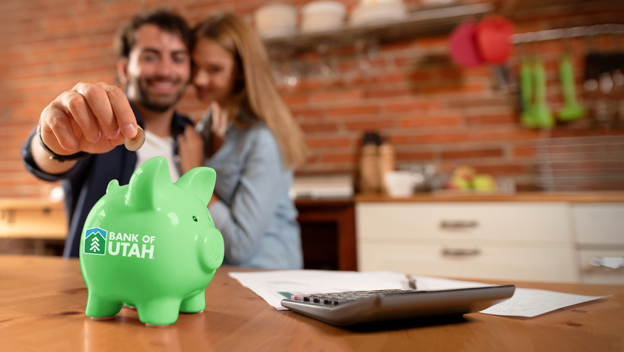 Happy couple in background reaches to put a coin into a green Bank of Utah piggy bank. 